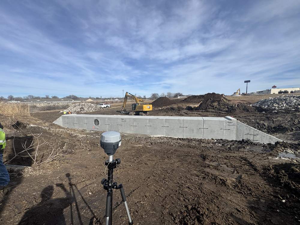 A GNSS survey receiver on a tripod on an active job site, with a precast concrete box culvert being set behind it and an excavator working beyond.